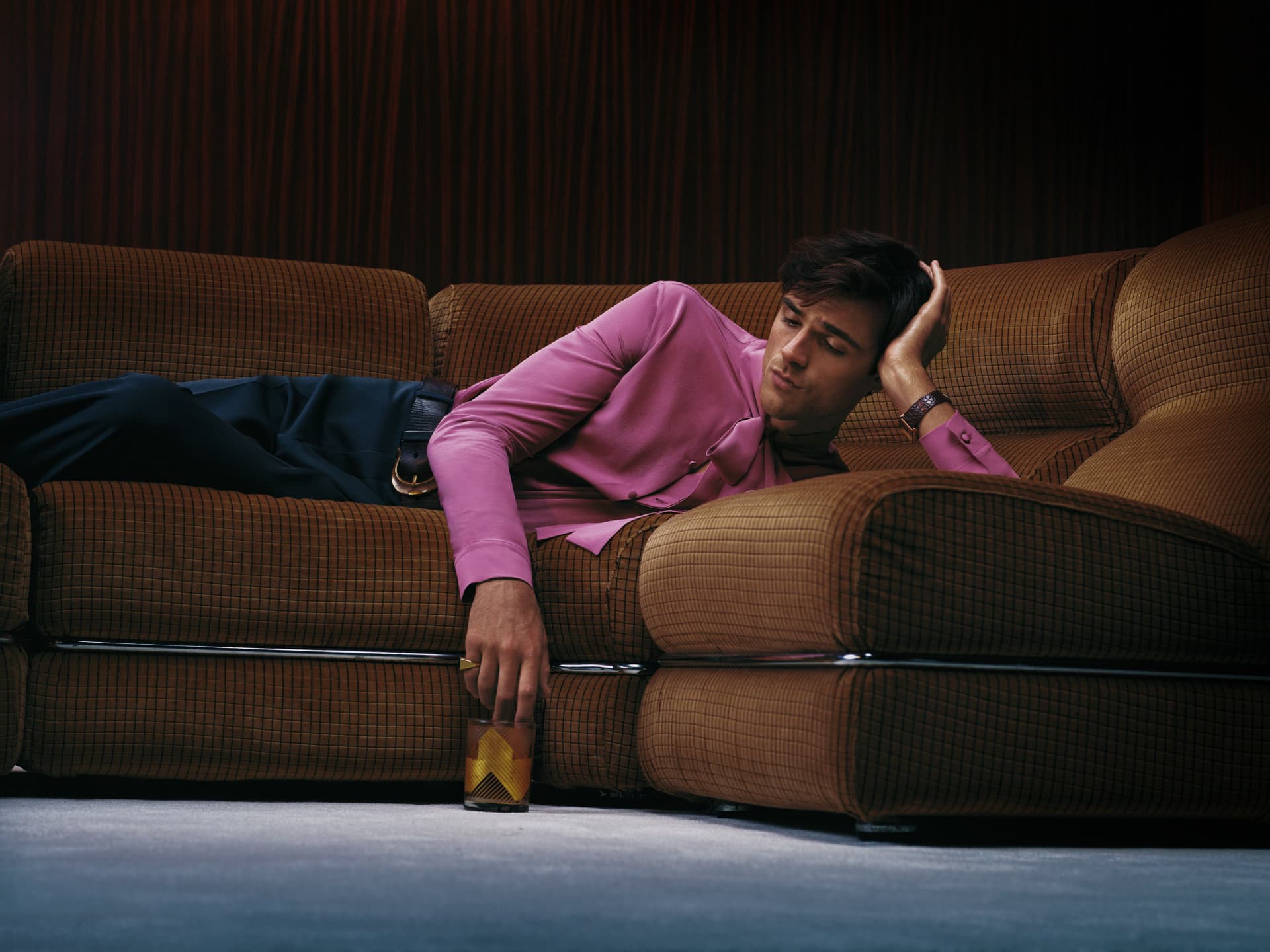 Jacob Elordi — reclining on a brown sofa in pink shirt holding a glass